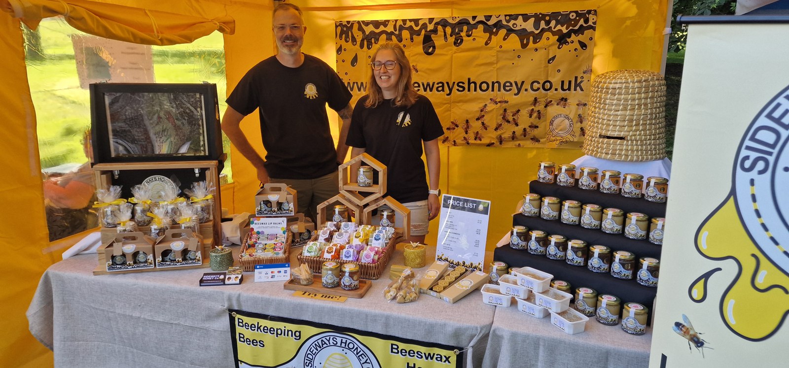 A smiling couple behind their Sideways Honey stall, displaying honey jars, beeswax products and gift sets at Christchurch Park Market